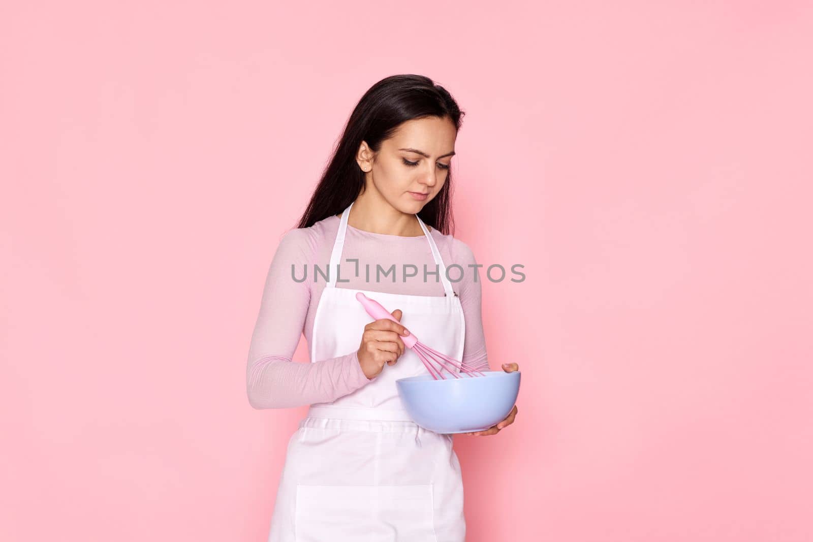 Pretty housekeeper baker woman holding whisk and bowl on pink background