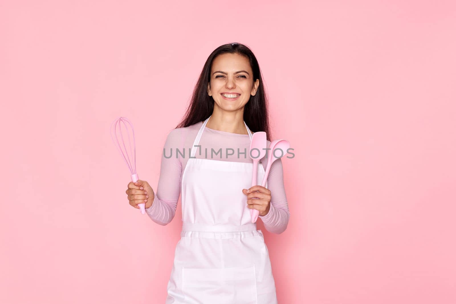 happy cooker woman in chef apron holding whisk, spoon and spatula on pink background