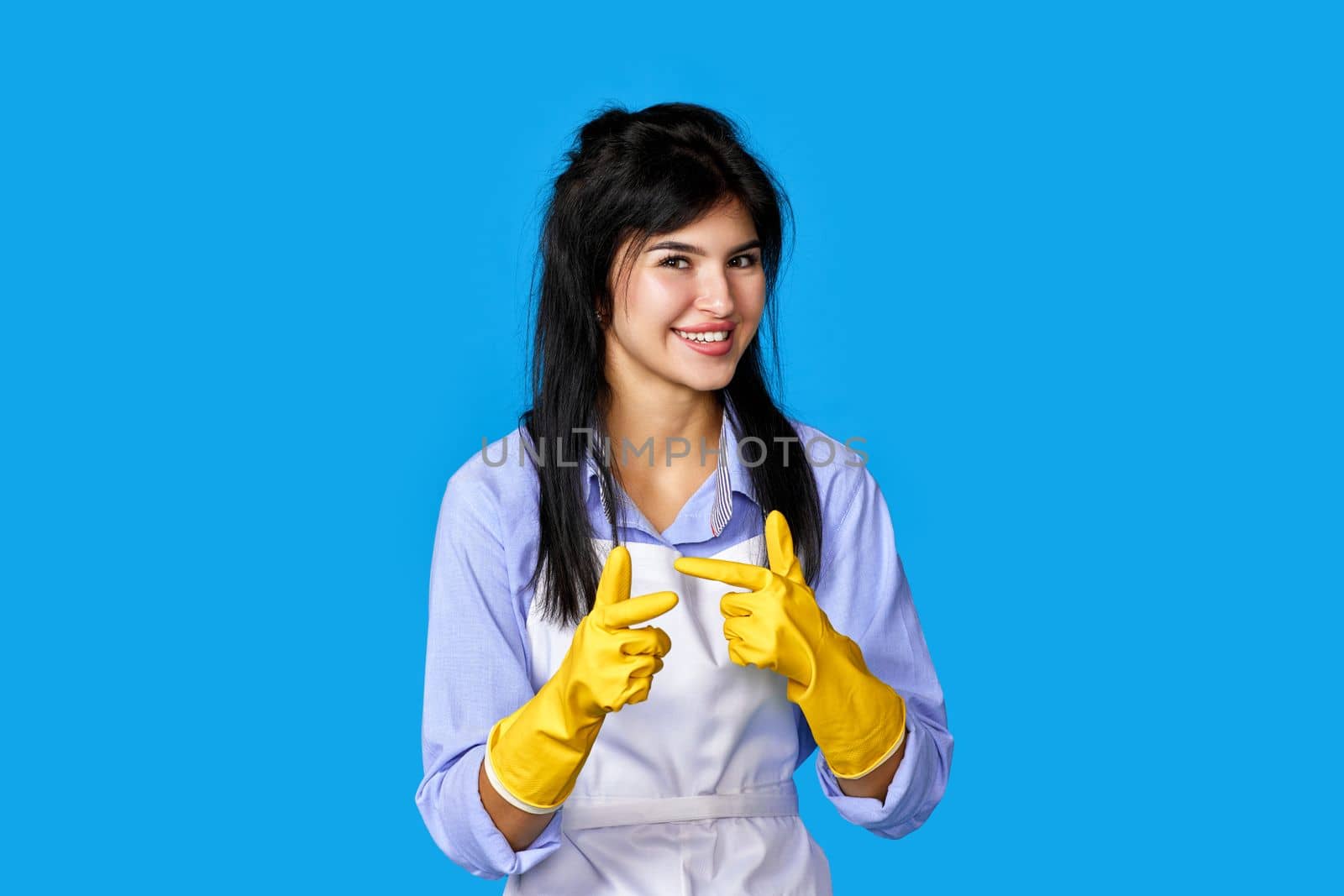 beautiful happy woman in yellow rubber gloves and cleaner apron pointing fingers to camera isolated on blue background. cleaning