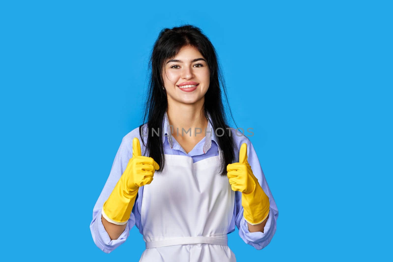 beautiful caucasian woman in yellow rubber gloves and cleaner apron showing ok sign isolated on blue background. cleaning