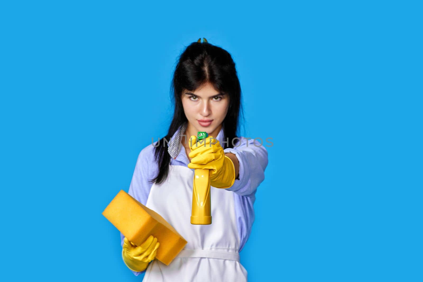 young confident woman in yellow rubber gloves and cleaner apron cleaning with sponge and detergent sprayer on blue background.