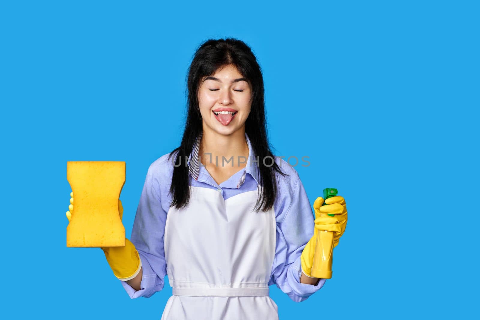 funny caucasian woman in yellow rubber gloves and cleaner apron with sponge and detergent sprayer isolated on blue background. cleaning