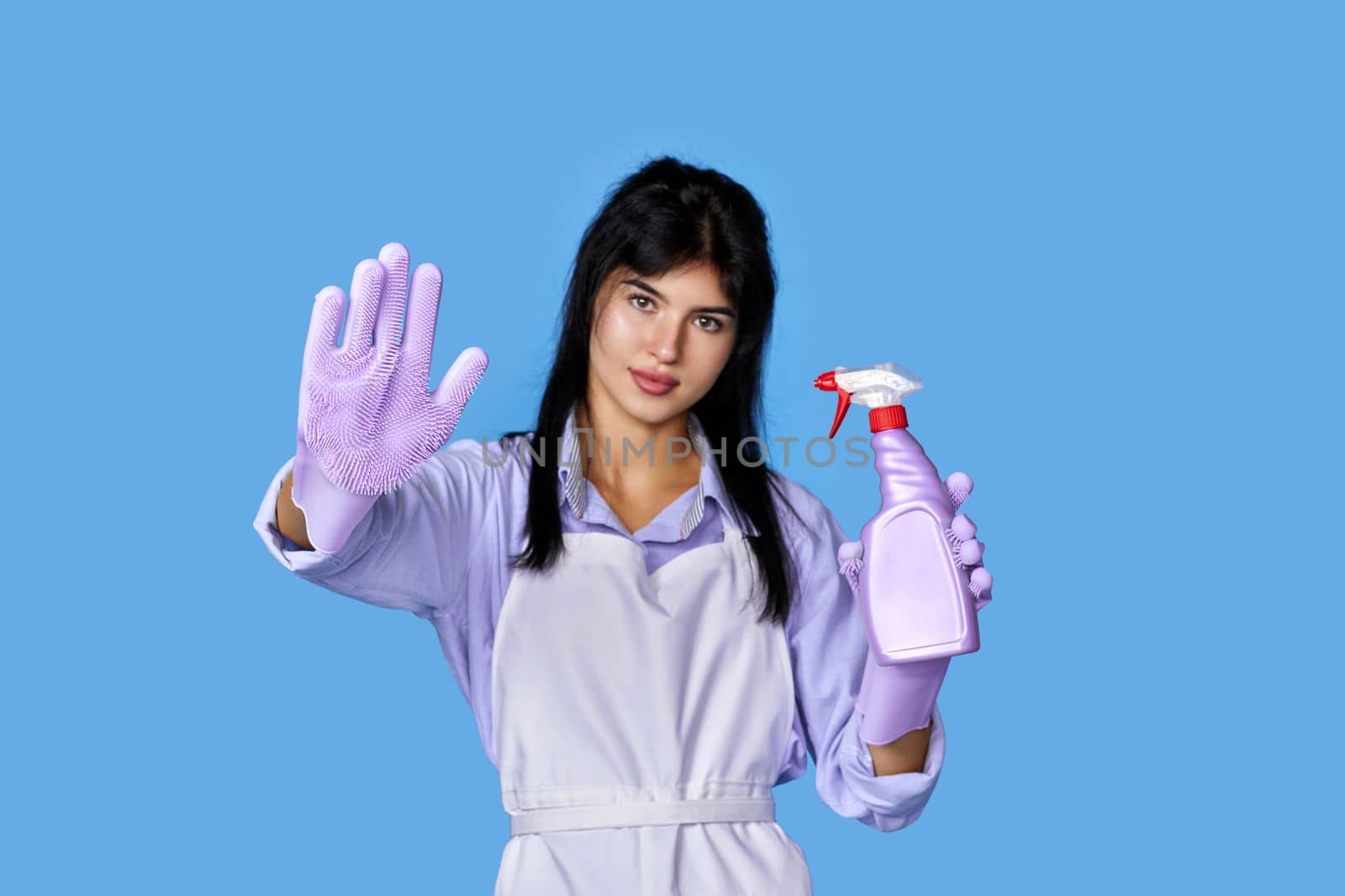 beautiful woman in rubber gloves and cleaner apron showing stop gesture isolated on blue background. focus on glove