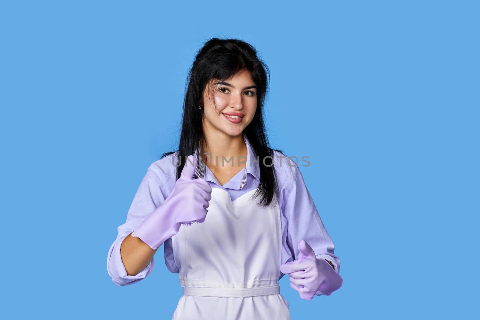 beautiful caucasian woman in violet rubber gloves and cleaner apron showing ok sign isolated on blue background. cleaning