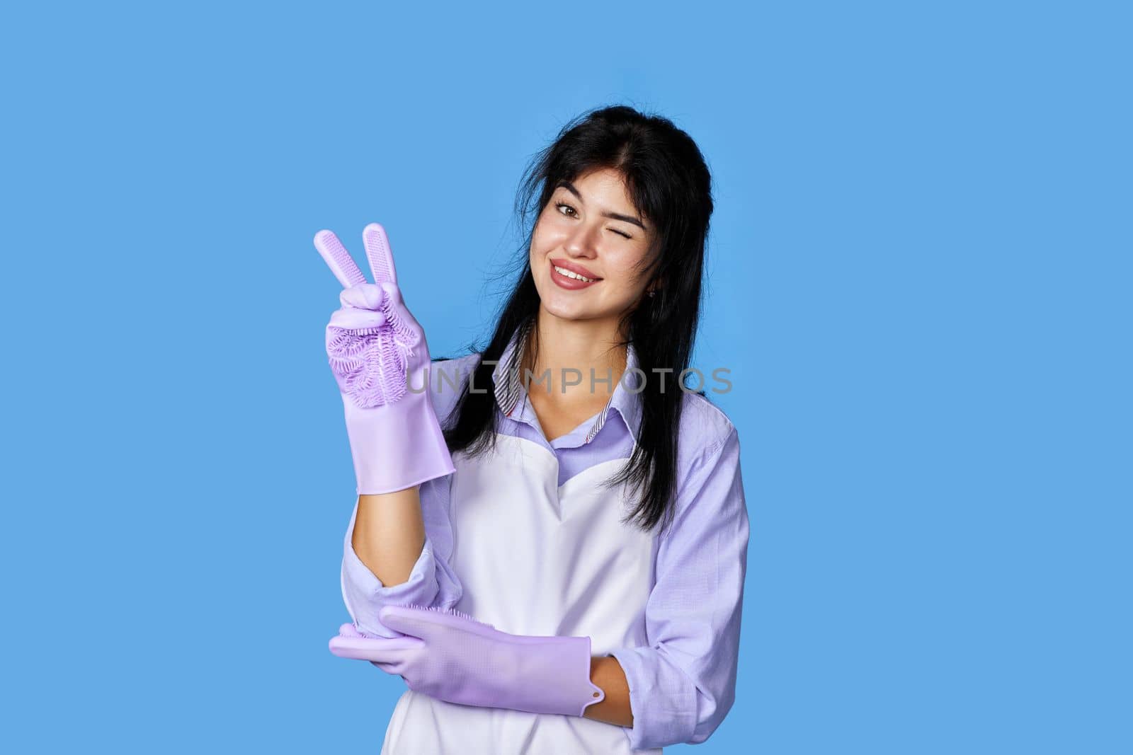 cheerful woman in rubber gloves and cleaner apron winking looking at the camera and showing victory gesture isolated on blue background. cleaning