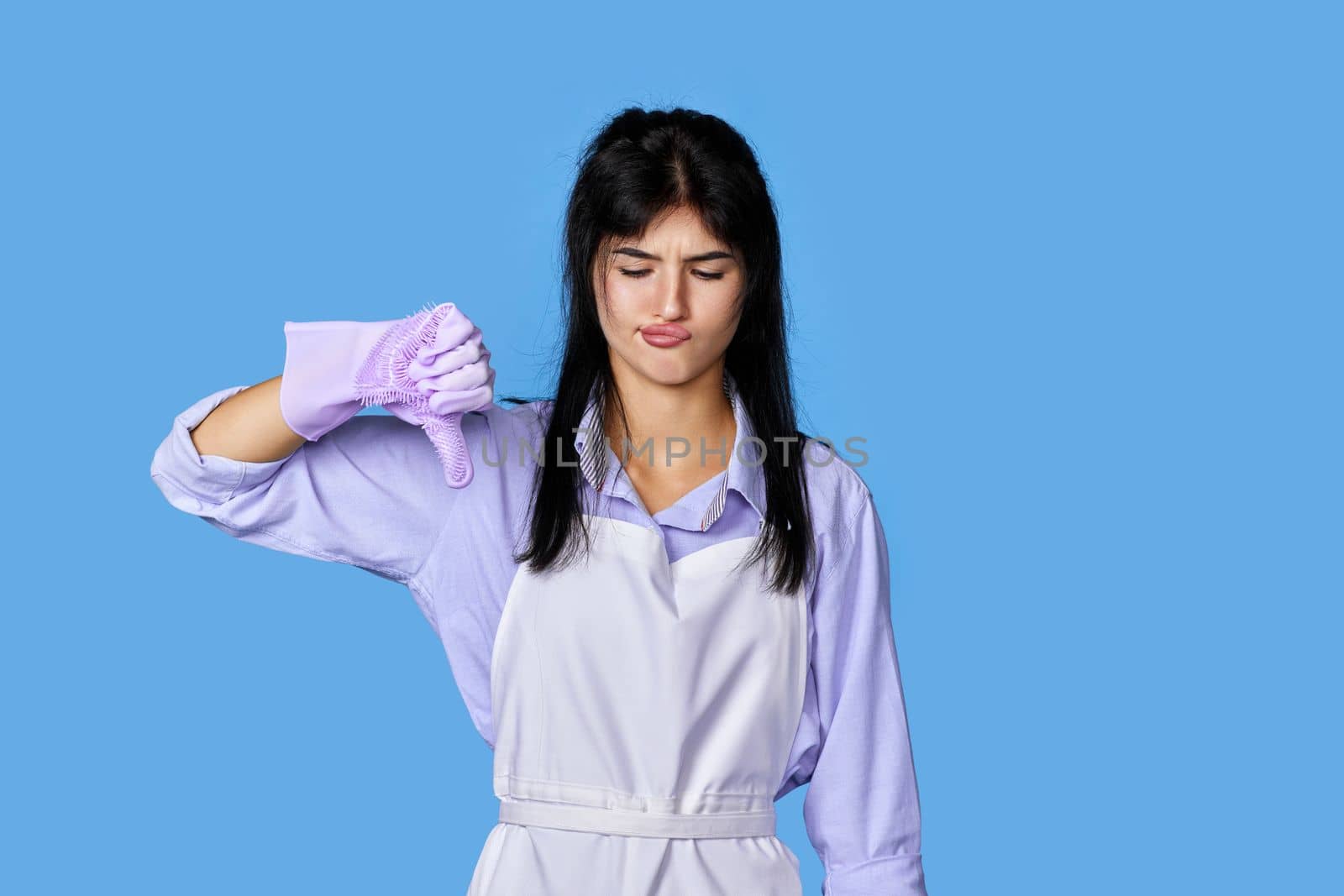 displeased woman in violet rubber gloves and cleaner apron showing thumbs down sign isolated on blue background. cleaning