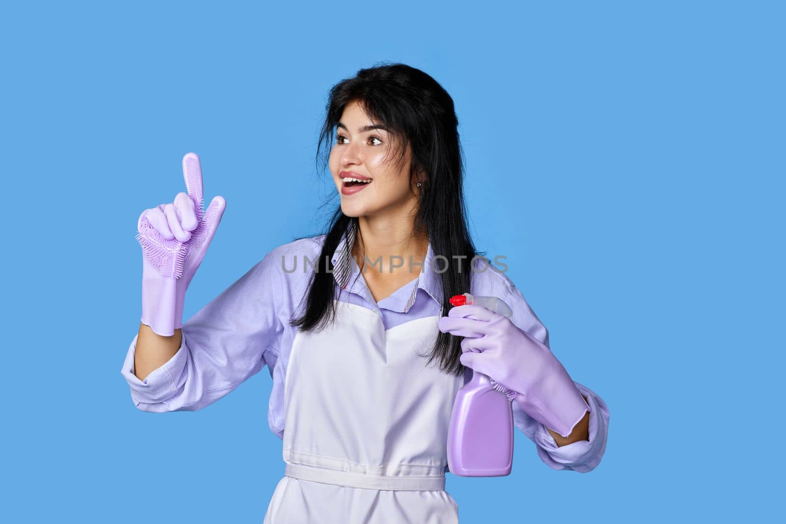 beautiful woman in violet rubber gloves and apron with detergent sprayer holding index finger up with new idea isolated on blue background. cleaning