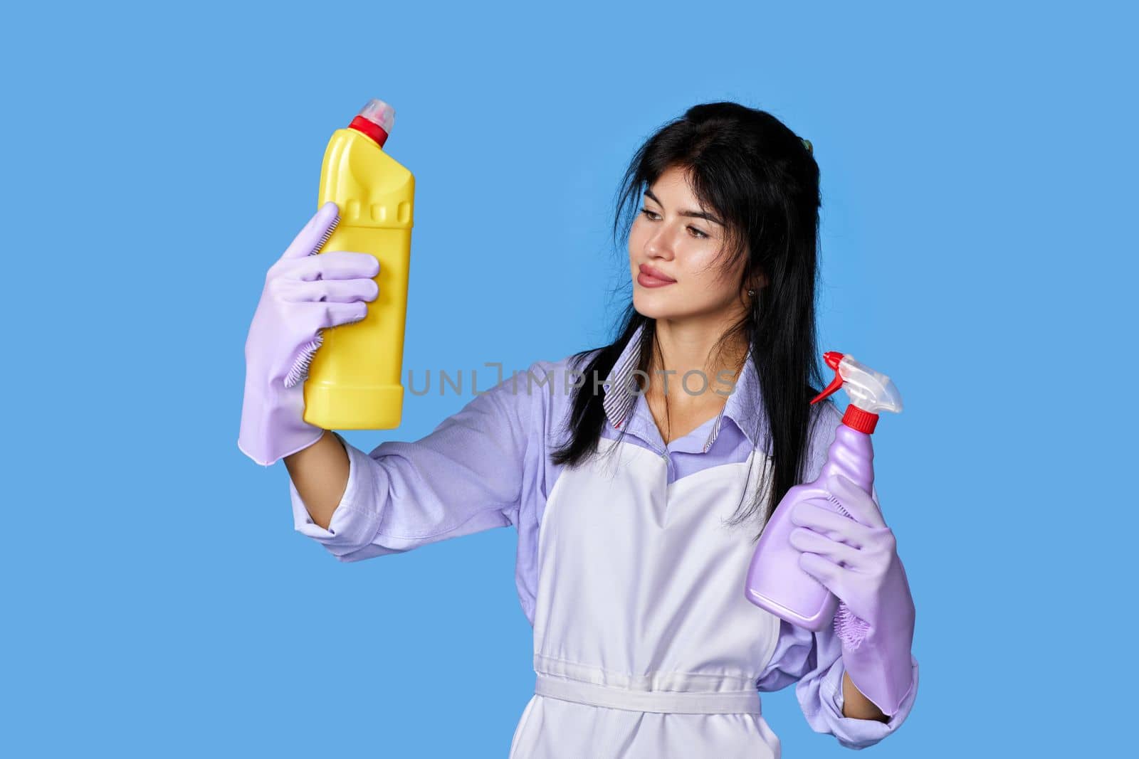 beautiful woman in violet rubber gloves and cleaner apron chooses between two cleaning products on blue background.