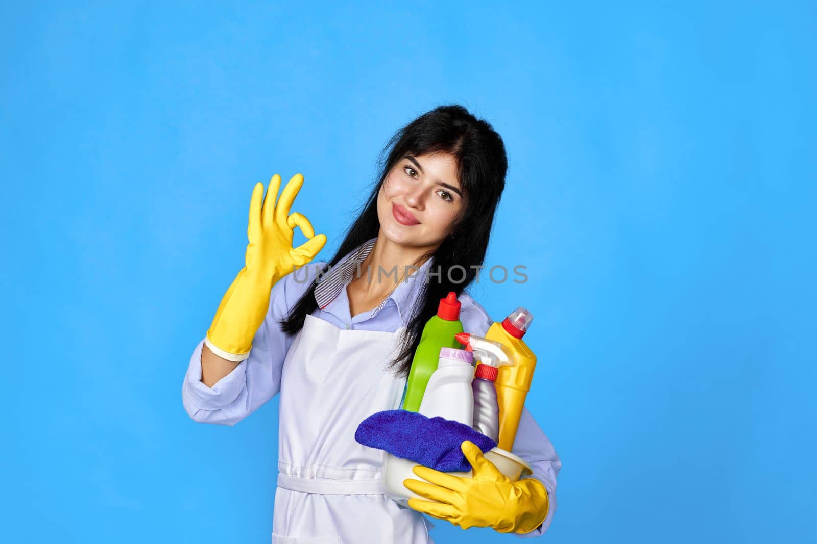 pretty woman in yellow rubber gloves and cleaner apron holding bucket of detergents and showing Ok gesture isolated on blue background.