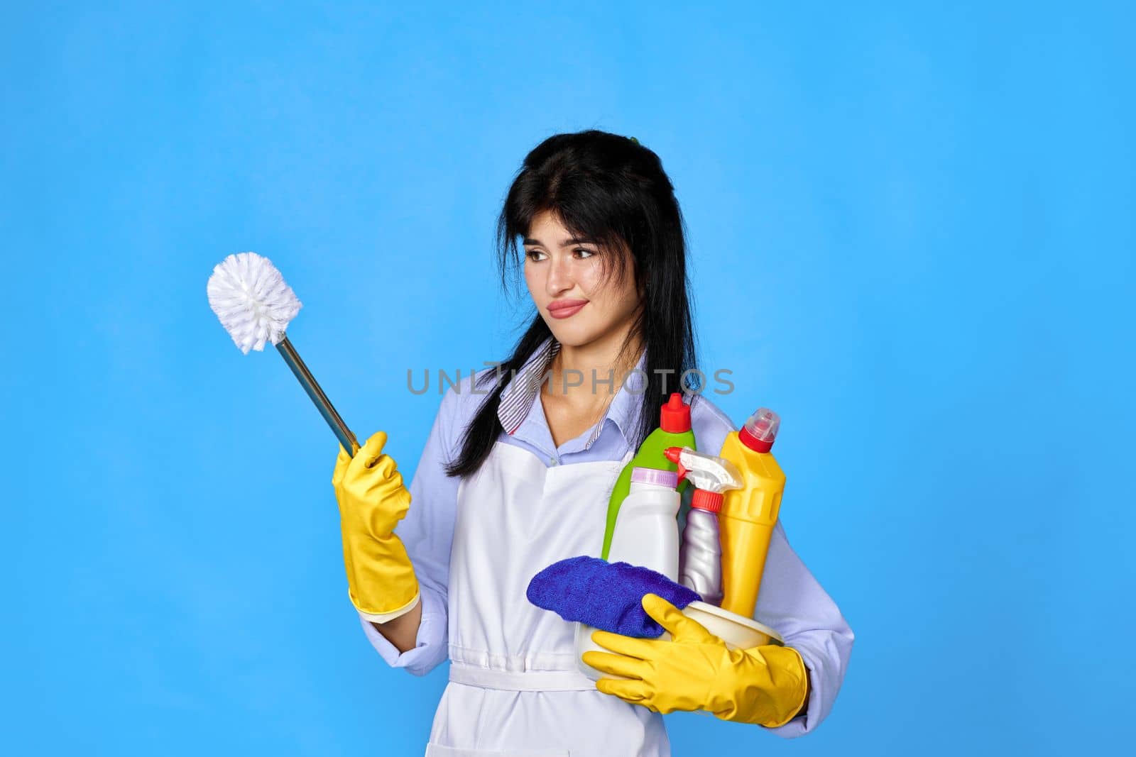 unhappy woman in yellow rubber gloves and cleaner apron holding bucket of detergents and toilet brush on blue background.