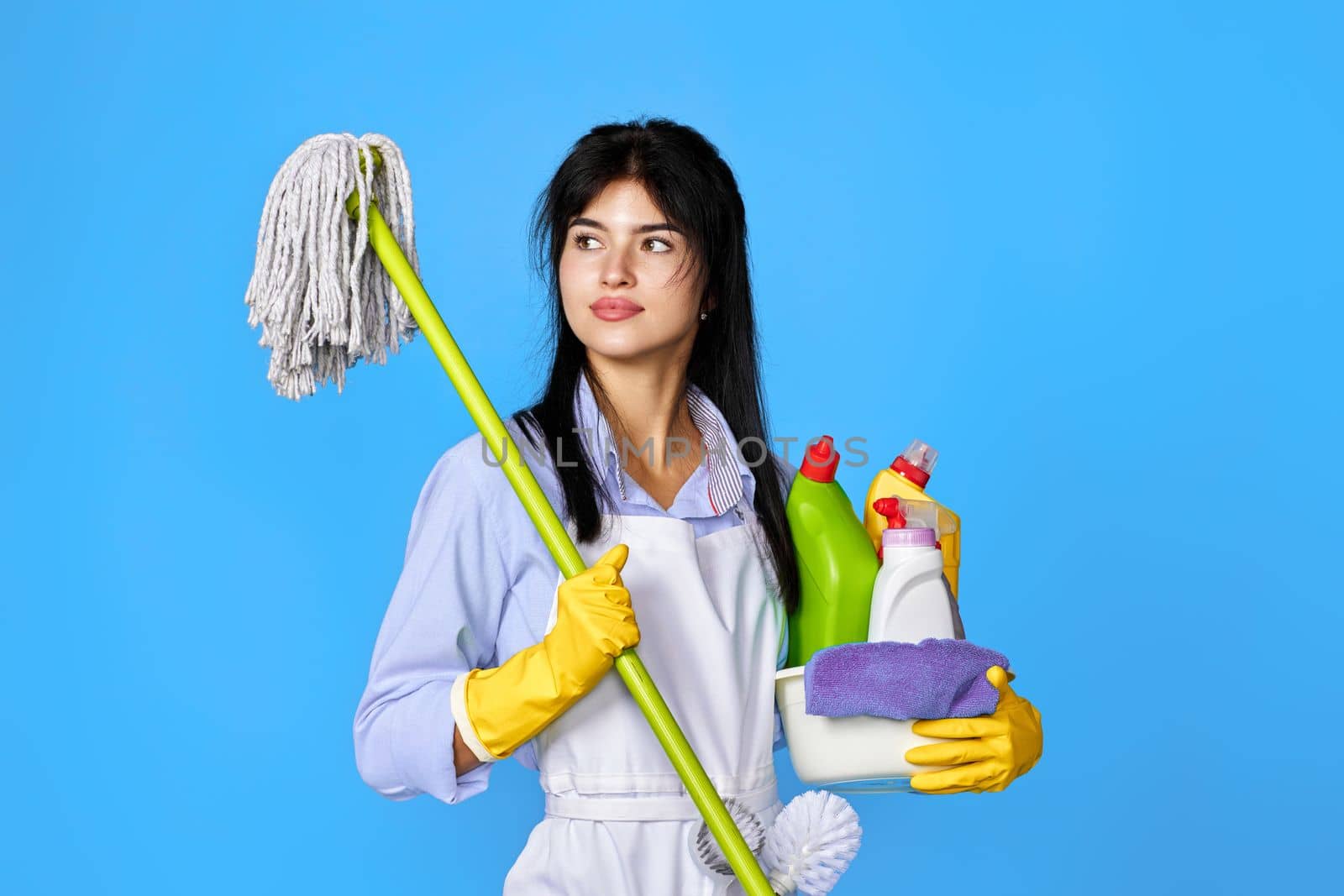 pretty woman in yellow rubber gloves and cleaner apron holding bucket of detergents and mop on blue background.