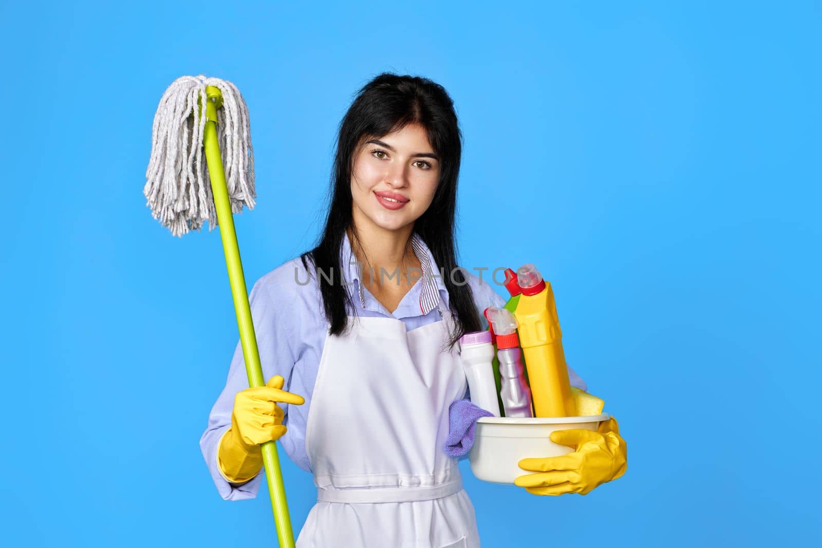 pretty woman in yellow rubber gloves and cleaner apron holding bucket of detergents and mop on blue background.