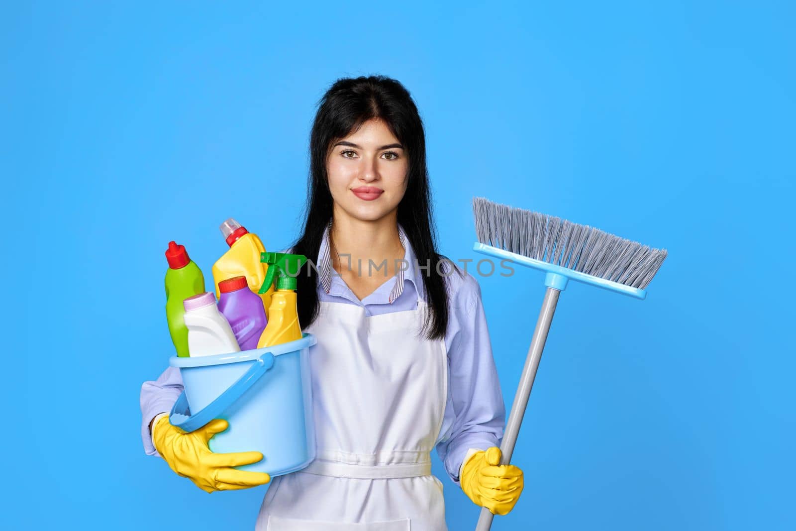 pretty woman in yellow rubber gloves and cleaner apron holding bucket of detergents and broom on blue background.