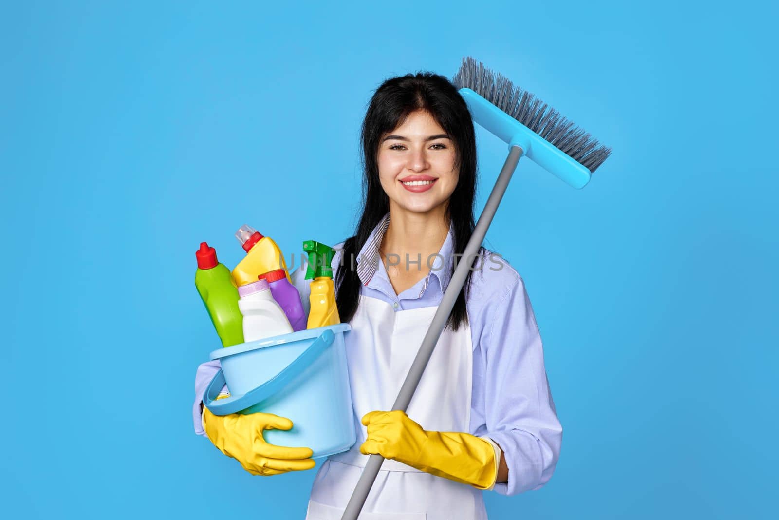 happy woman in gloves and cleaner apron holding bucket of detergents and broom on blue background.