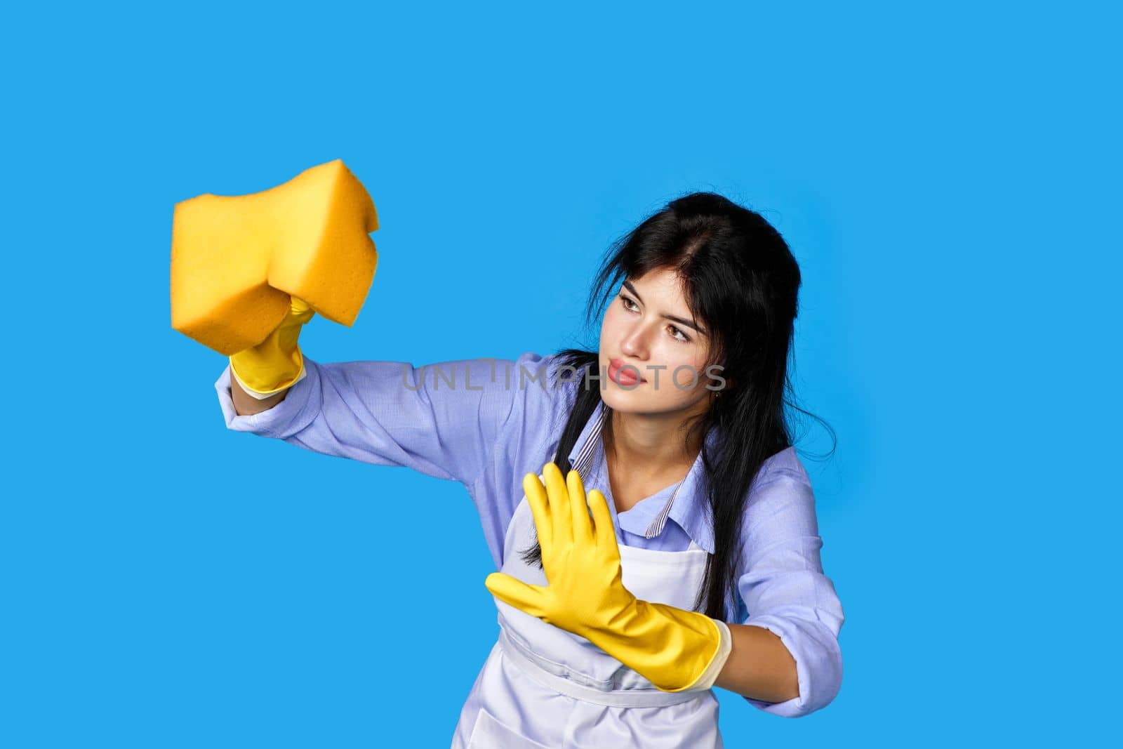 young attentive woman in rubber gloves and cleaner apron cleaning with yellow sponge on blue background.