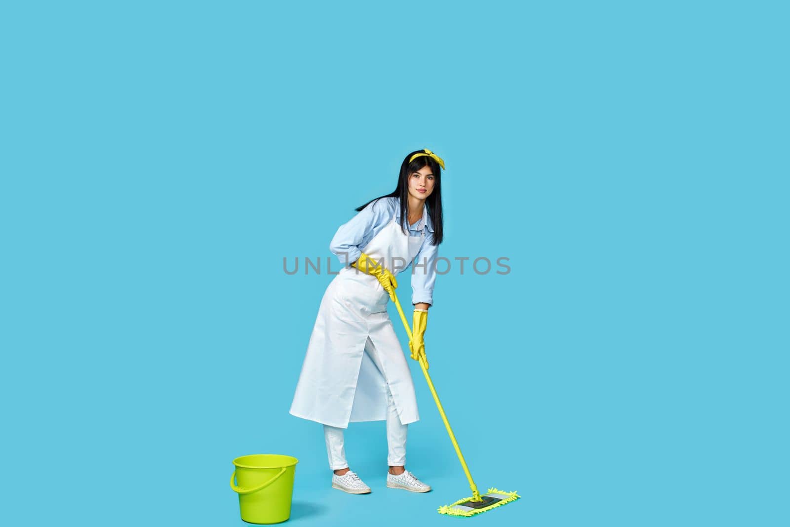 woman in rubber gloves and cleaner apron washing floor with mop on blue background. Full length