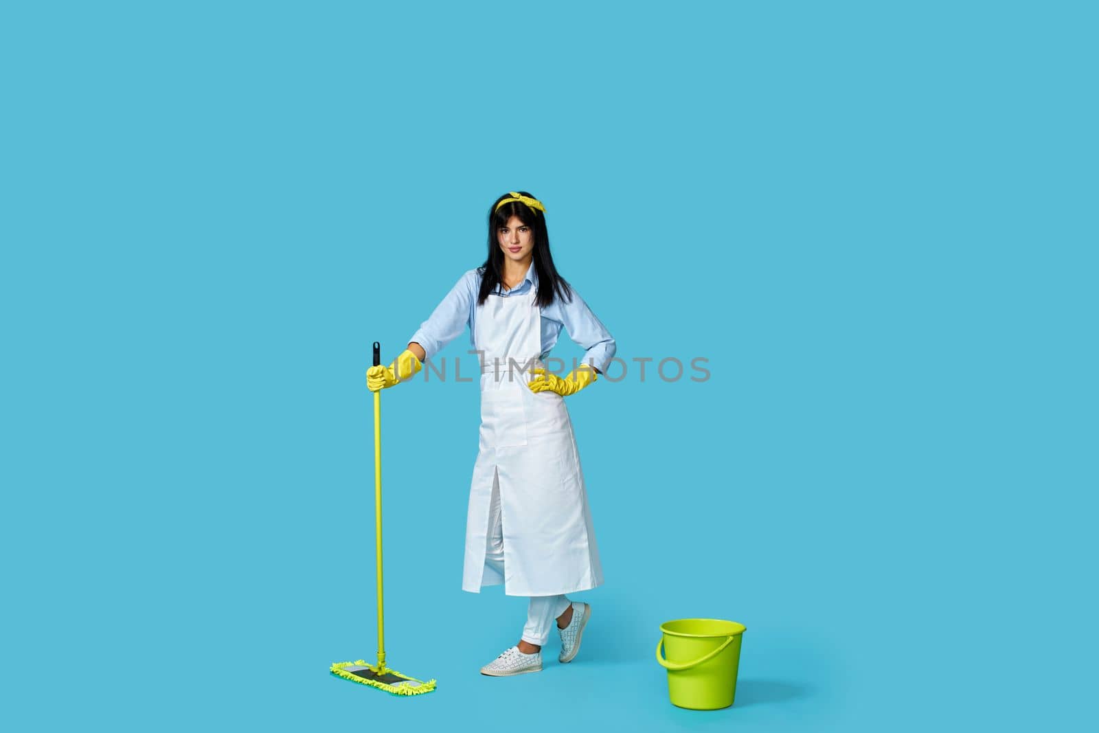 professional cleaner girl in rubber gloves and cleaner apron washing floor with mop on blue background. Full length