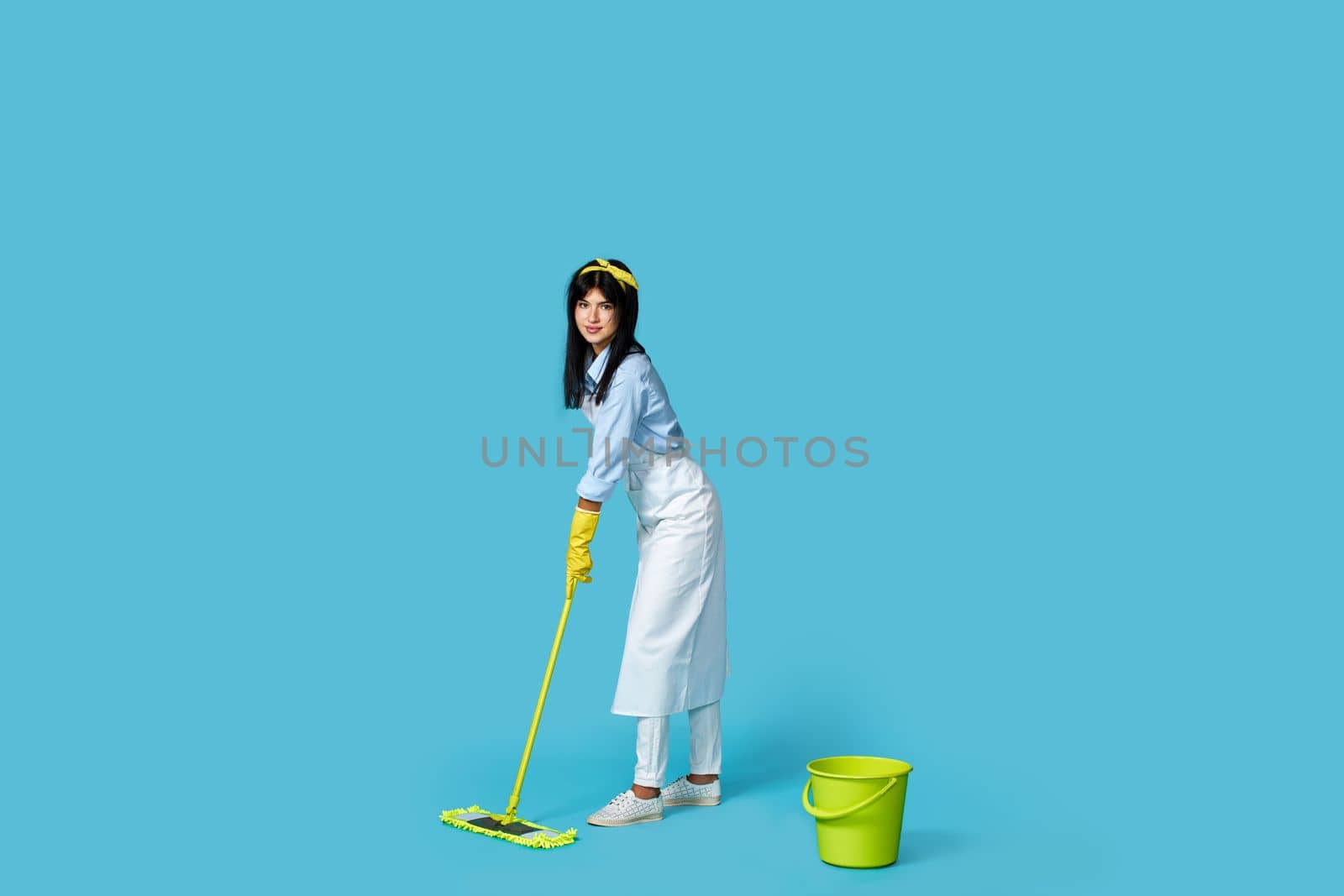 cheerful woman in rubber gloves and cleaner apron washing floor with mop on blue background. Full length