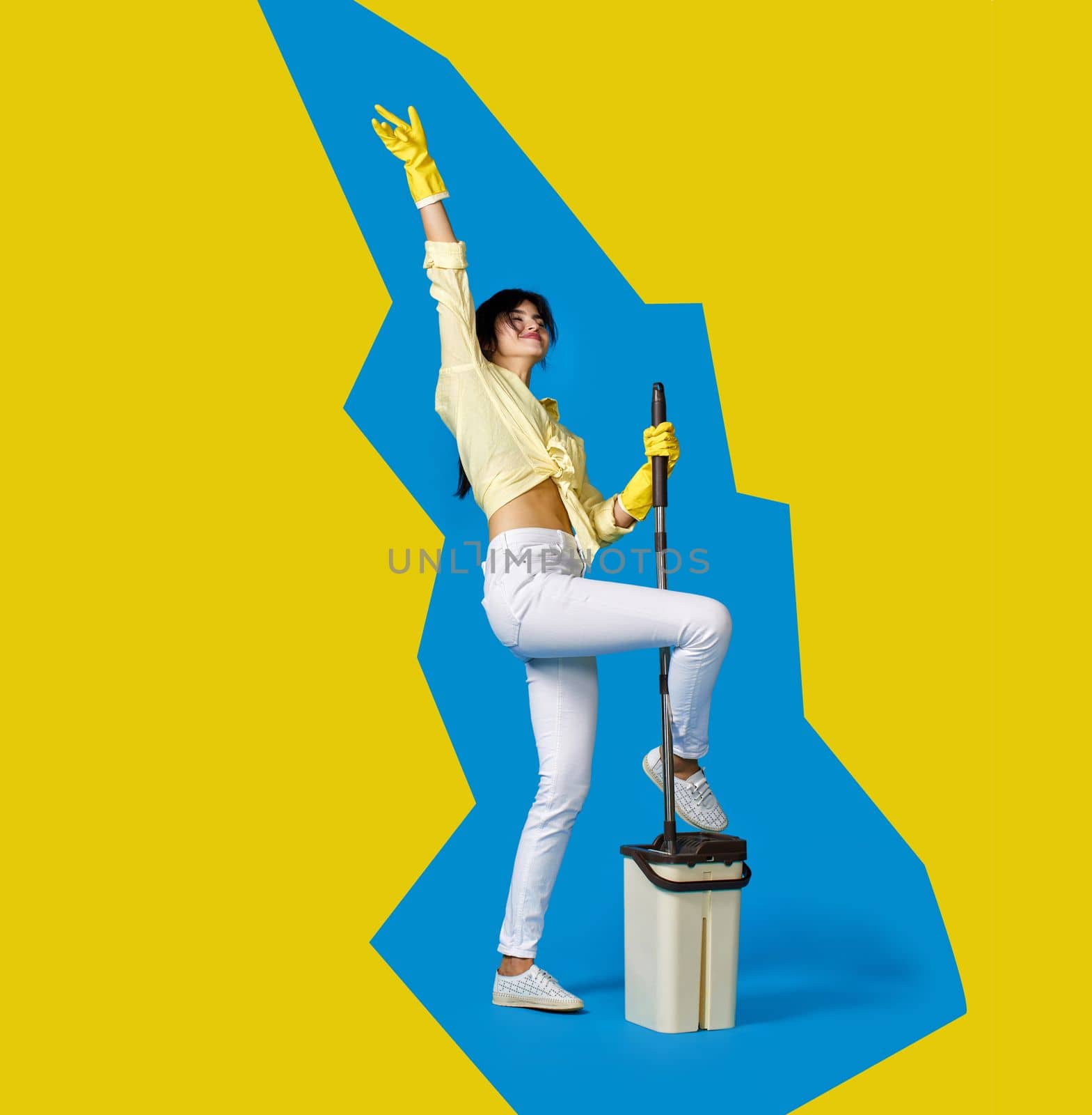 funny housewife girl in rubber gloves washing floor with mop and dancing on blue and yellow background. Full length