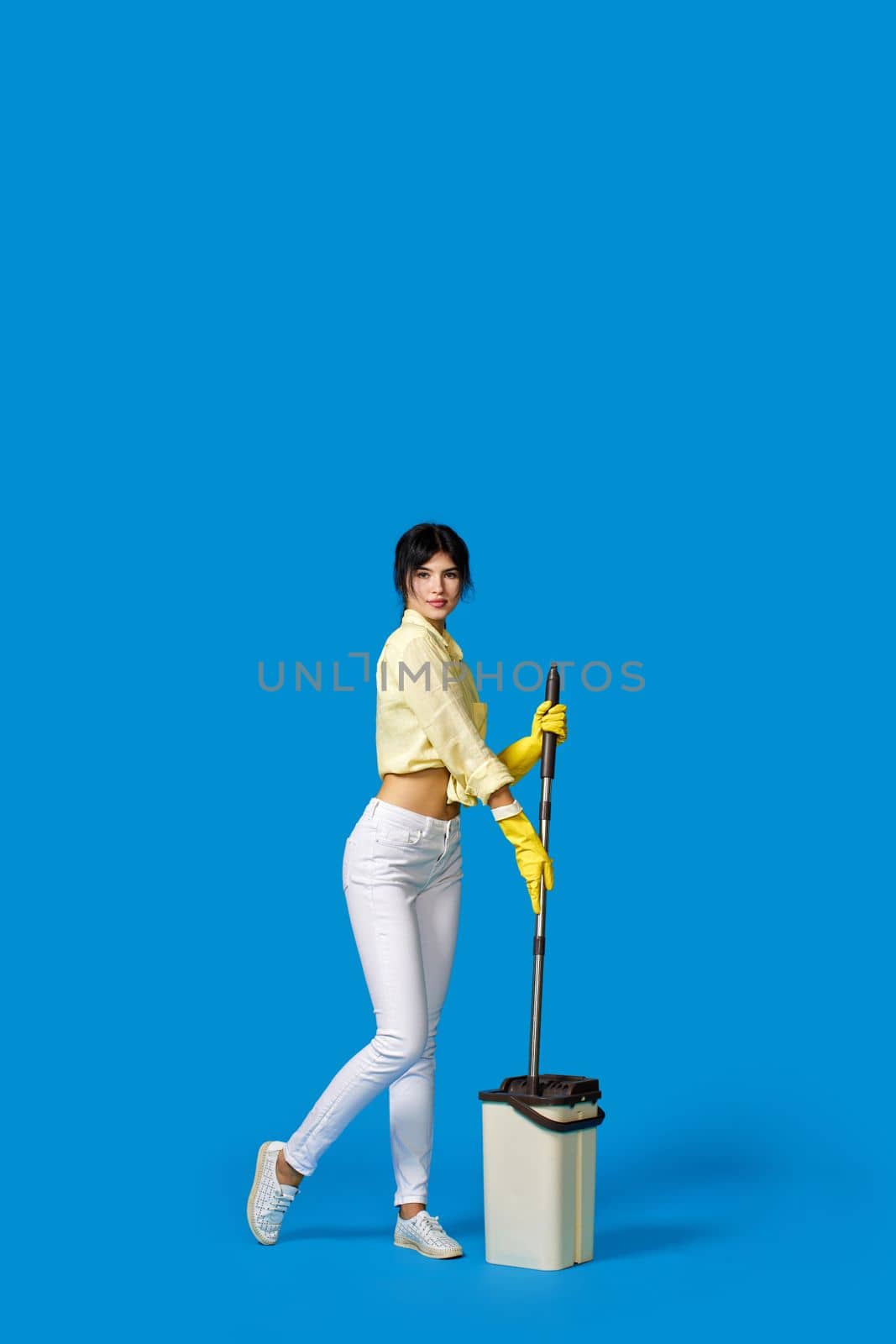 beautiful housewife girl in rubber gloves washing floor with mop on blue background. Full length