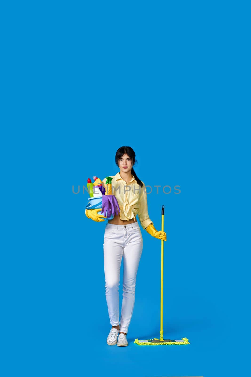 concenteated serious girl in rubber gloves housekeeping and cleaning with mop and holding bucket of detergents on blue background. Full length