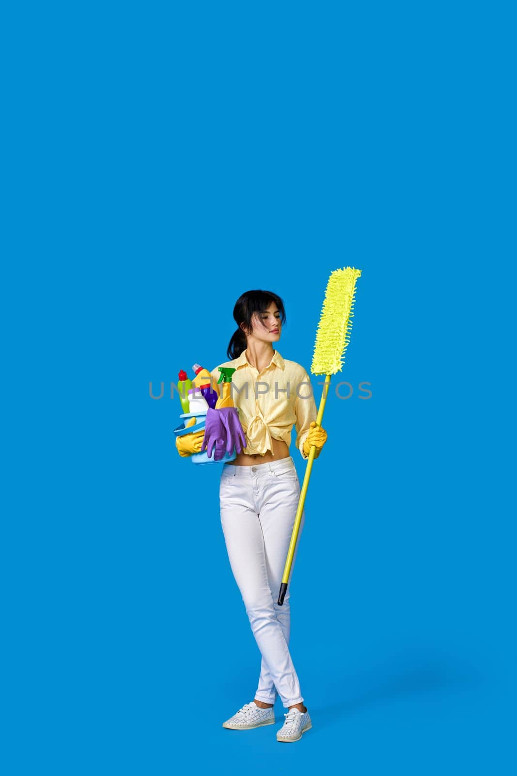 caucasian housewife girl in rubber gloves with mop and holding bucket of detergents on blue background. Full length