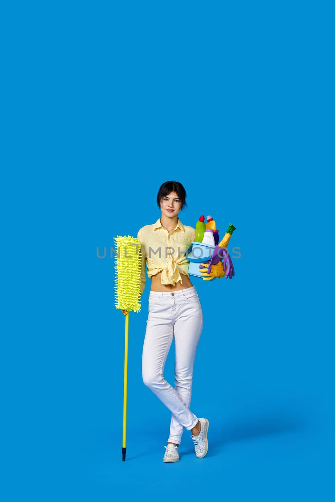 concenteated serious housewife girl in rubber gloves housekeeping and cleaning with mop and holding bucket of detergents on blue background. Full length