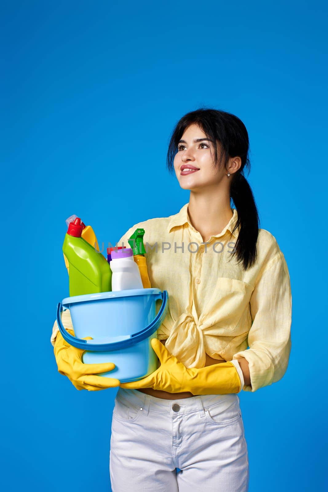 caucasian brunette woman in yellow gloves holding bucket with cleaning supplies isolated on blue background.