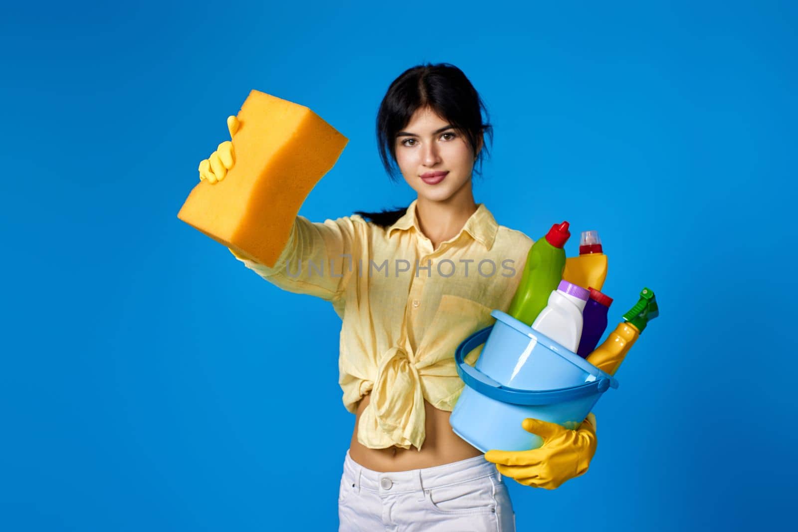 brunette woman in yellow gloves housekeeping and cleaning with sponge and bucket with cleaning supplies on blue background. focus on sponge