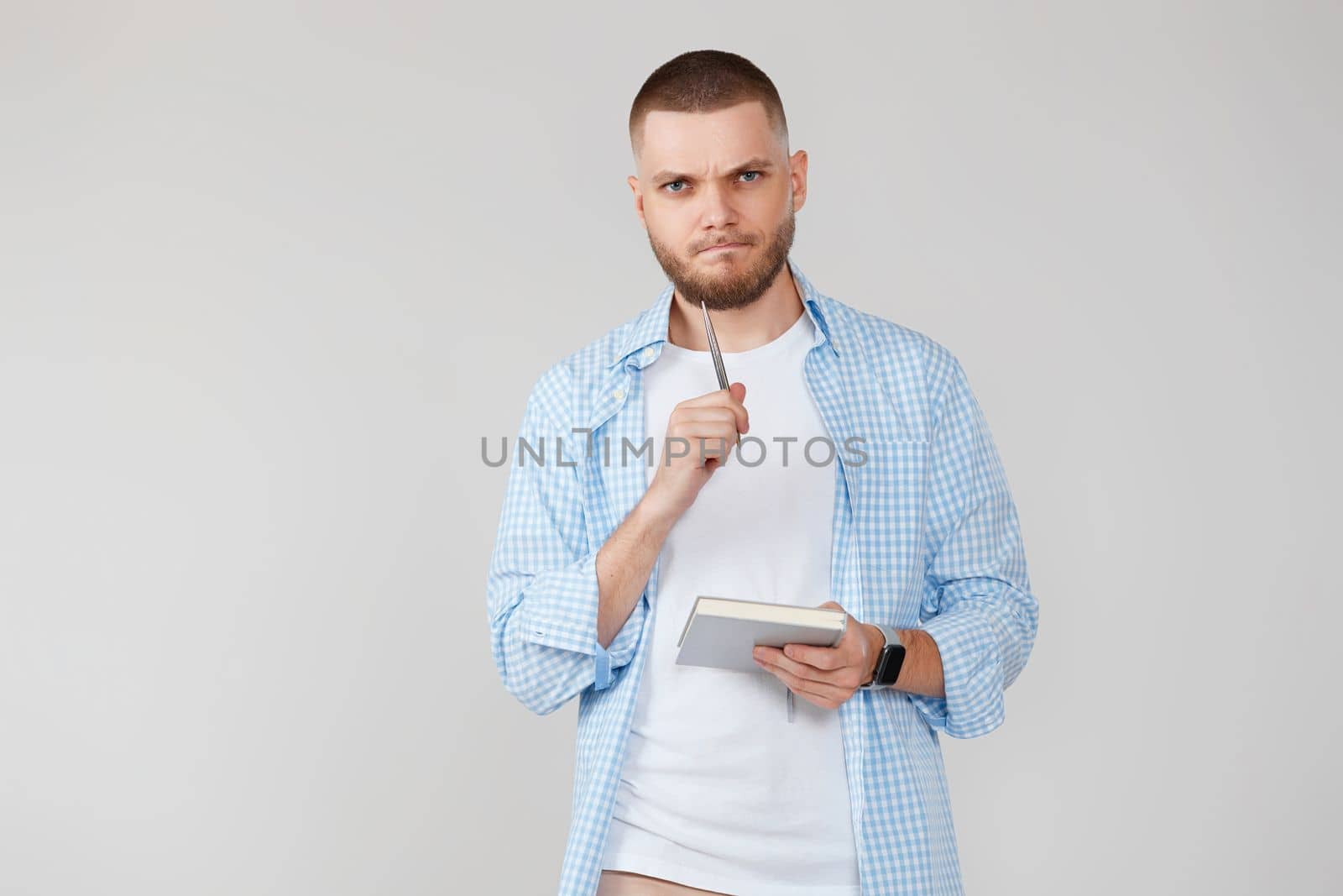 serious handsome man in casual blue shirt holding notebook and thoughtful about confusing idea on gray background