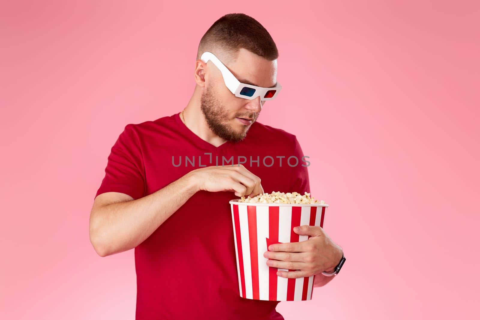 Young caucasian man in 3d glasses eating popcorn on pink background