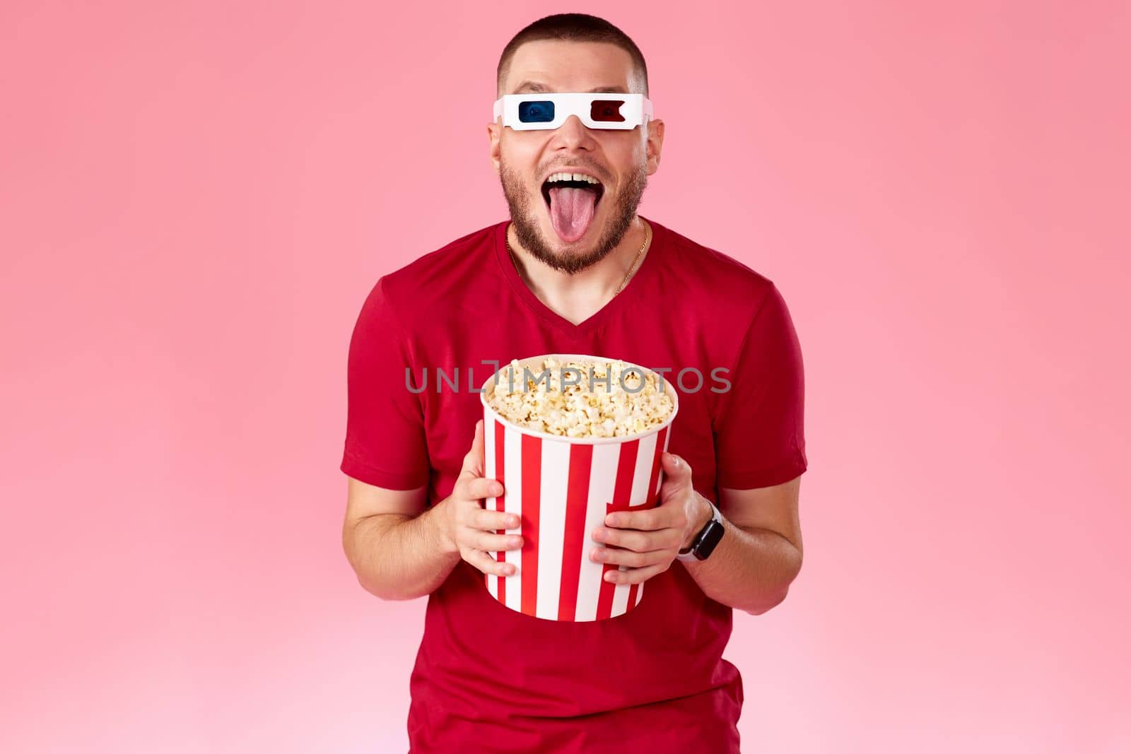 Young caucasian man in 3d glasses eating popcorn on pink background
