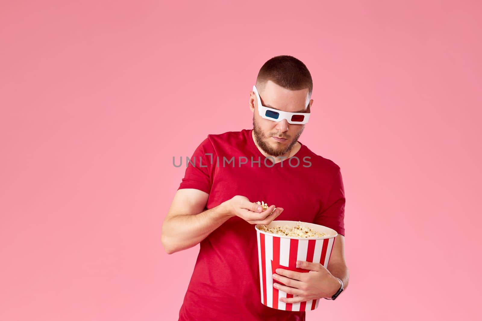 Young caucasian man in 3d glasses eating popcorn on pink background