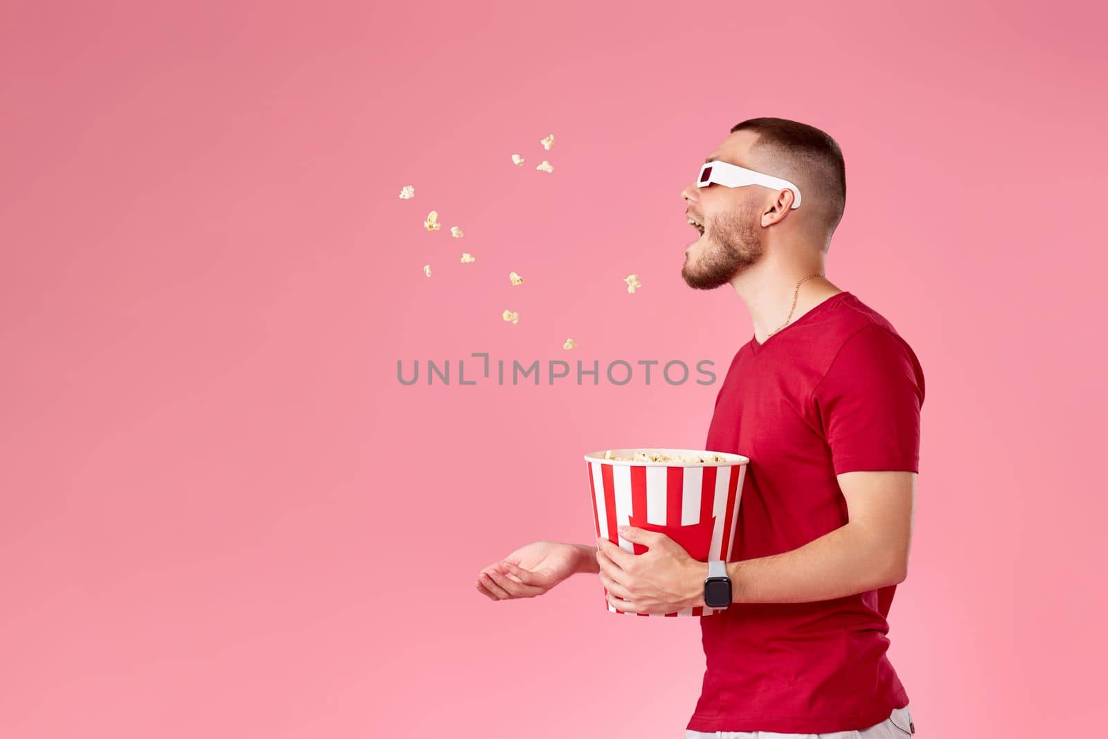 young man in 3d glasses and red t-shirt throwing up and eating popcorn on pink background. copy space