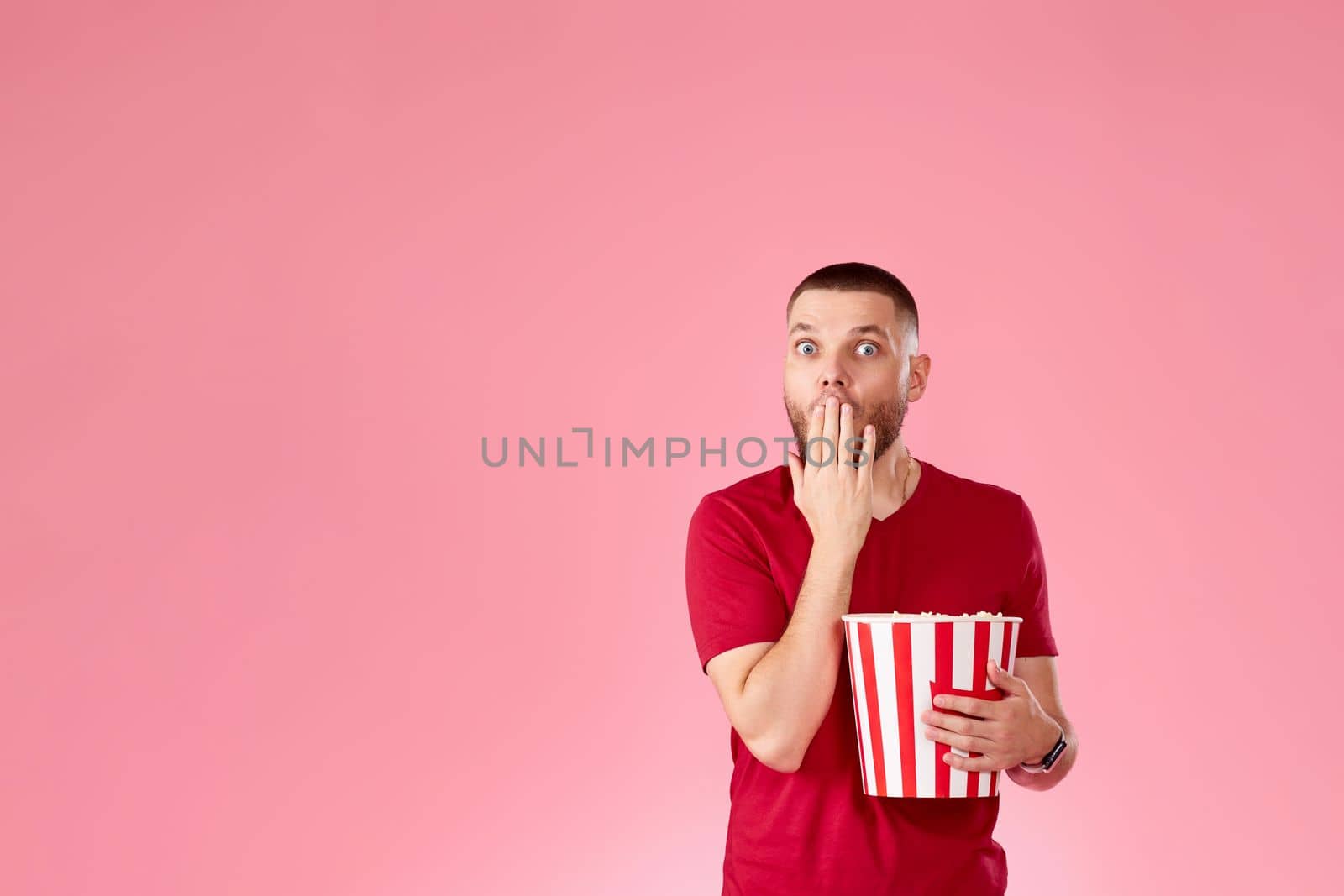 Shocked scared young caucasian man eating popcorn on pink background. copy space