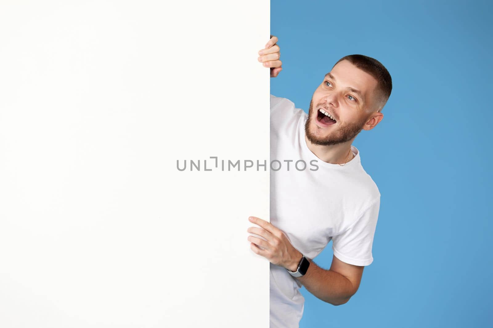 Portrait of friendly man in casual t-shirt peeking out from behind advertisement whiteboard with empty copy space on blue background