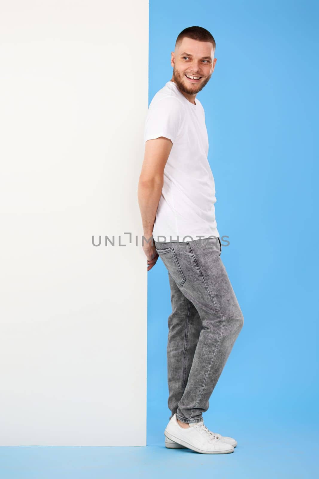Portrait of friendly guy in casual t-shirt leaning on empty white placard for advertisement on blue background