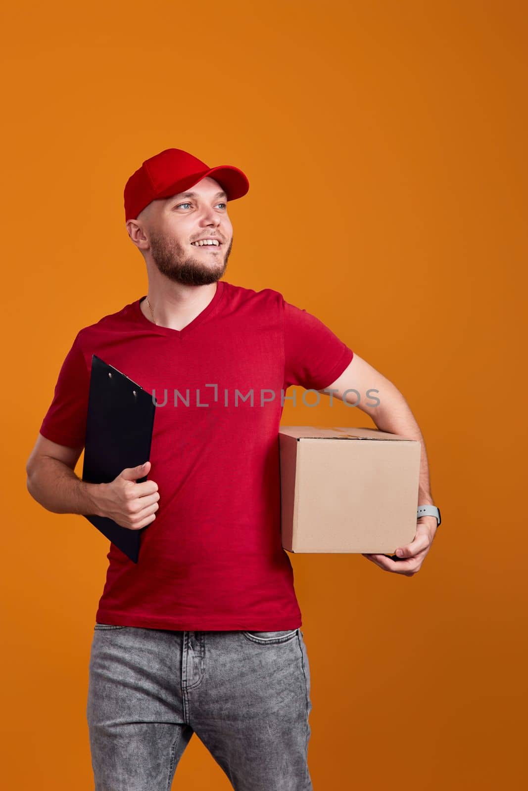 Delivery man in red cap, t-shirt holding clipboard and empty cardboard box isolated on orange background