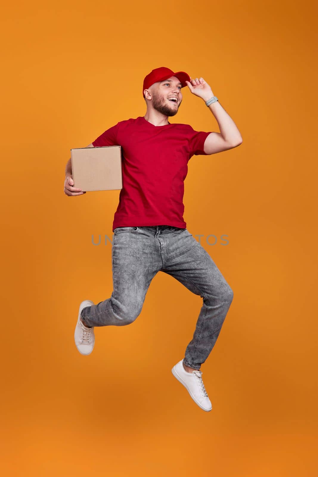 Delivery man in red cap, t-shirt holding cardboard box and jumping isolated on orange background