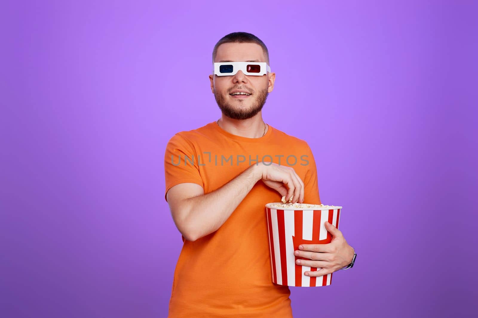happy caucasian man in 3d glasses eating popcorn on violet background