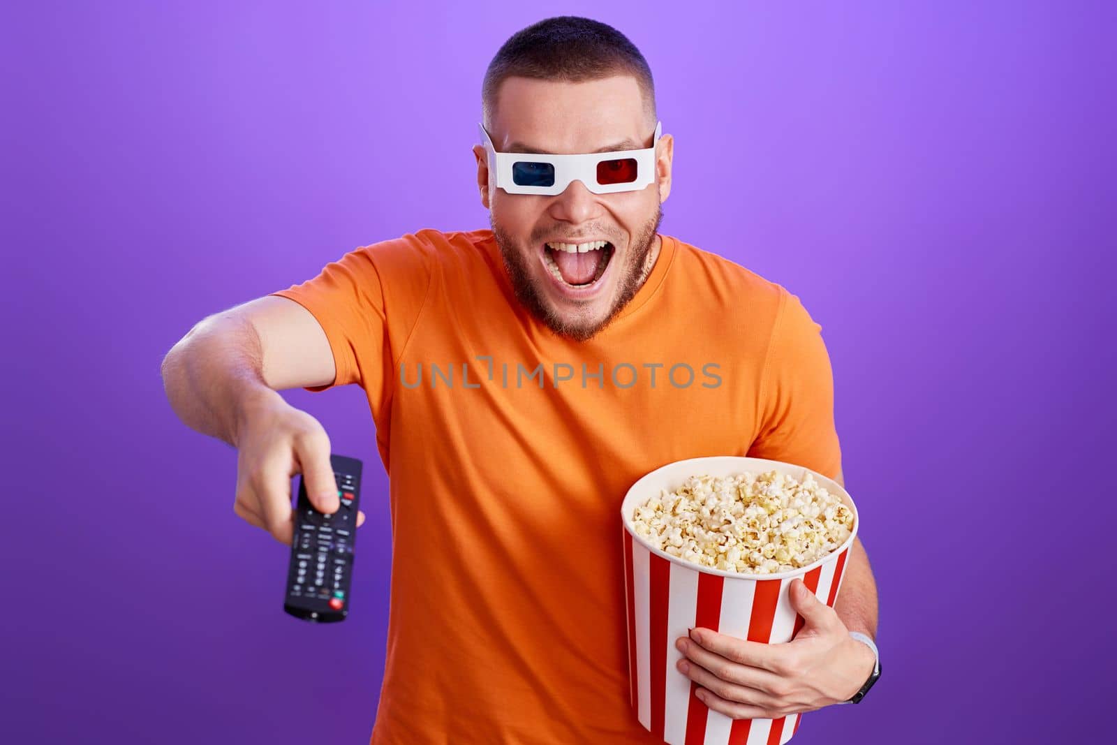 surprised man in 3d glasses using remote controller watching movie and holding big bucket of popcorns on purple background