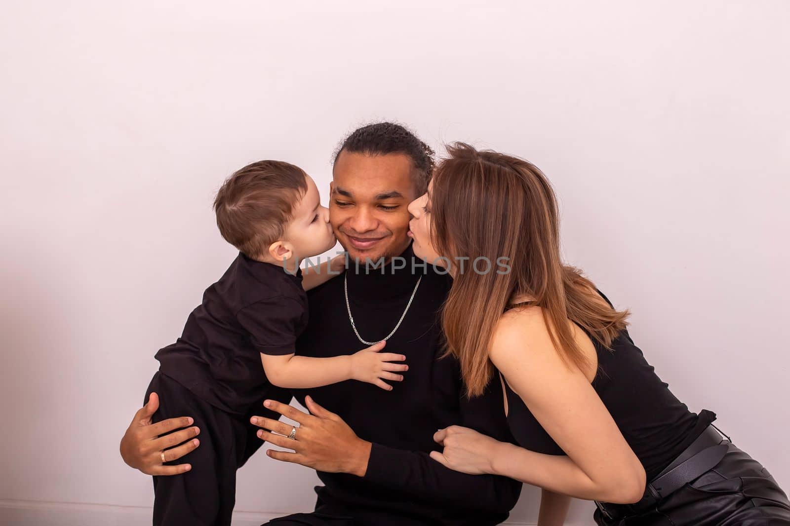 a happy family, a woman and a little boy, in black clothes, kiss a stylish young man on the cheeks from both sides, sit near a light wall. Copy space. Close up