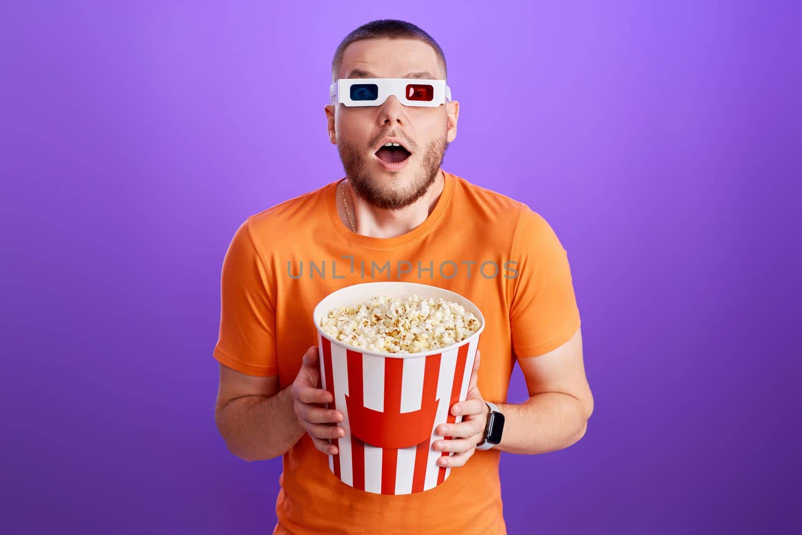amazed caucasian man in 3d glasses holding a big bucket of popcorns on purple background