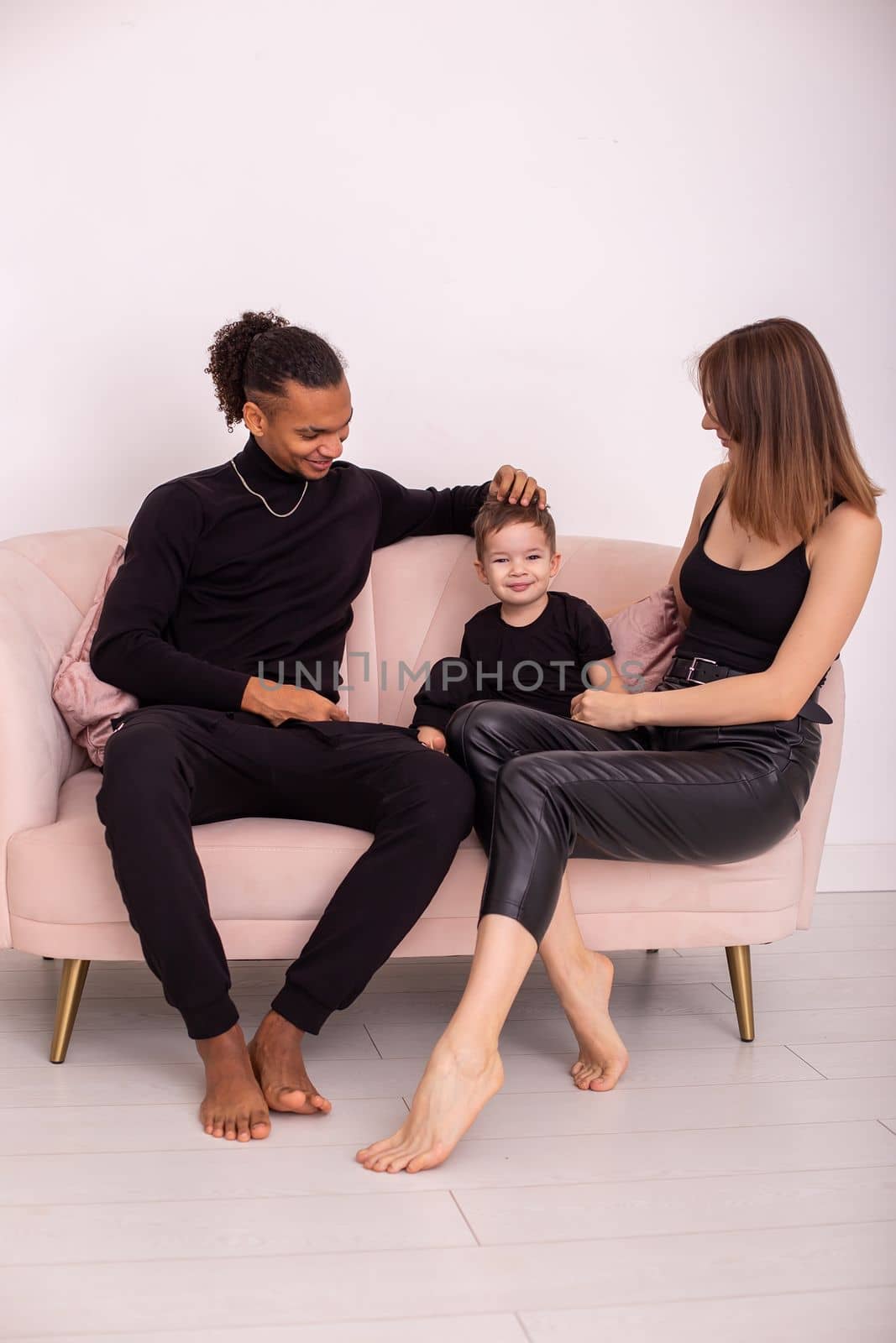 Happy multiracial family with little boy, in black clothes, sits in the light living room on light pink sofa, communicate. Vertical. Copy space