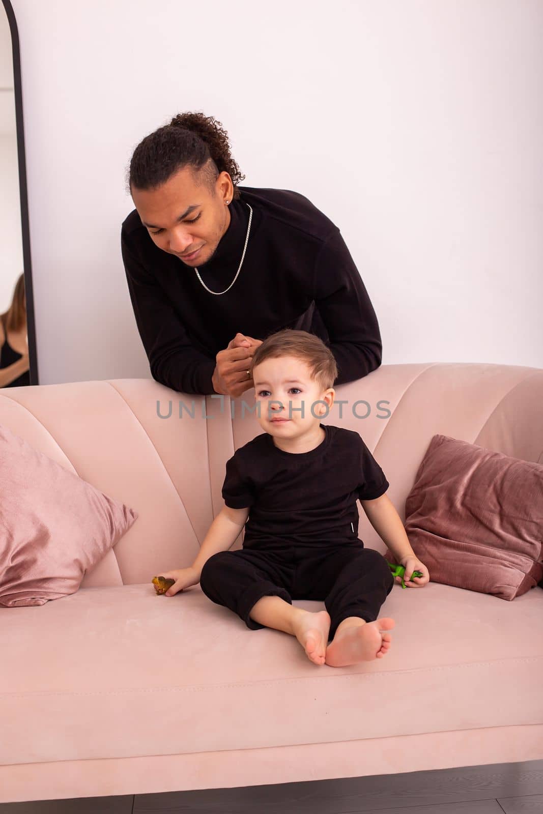 Smiling father and little son, in black clothes, sitting on ligh pink sofa in the room, light interior. Vertical. Copy space