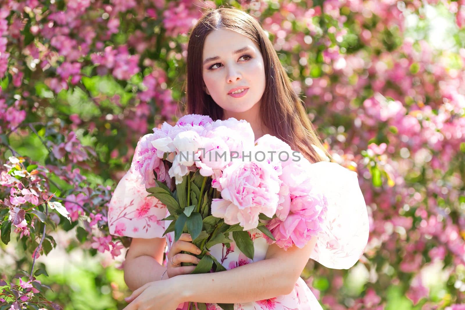 A portrait of a beautiful girl holding a large bouquet of pink peonies in her hands stands in a blooming garden on a sunny day. Copy space
