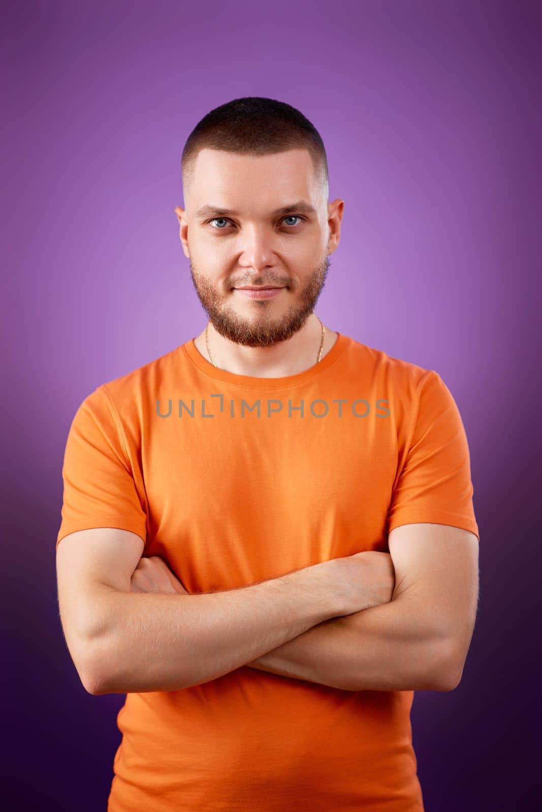 smiling young guy in orange t-shirt posing with crossed arms and looking at the camera on purple studio background