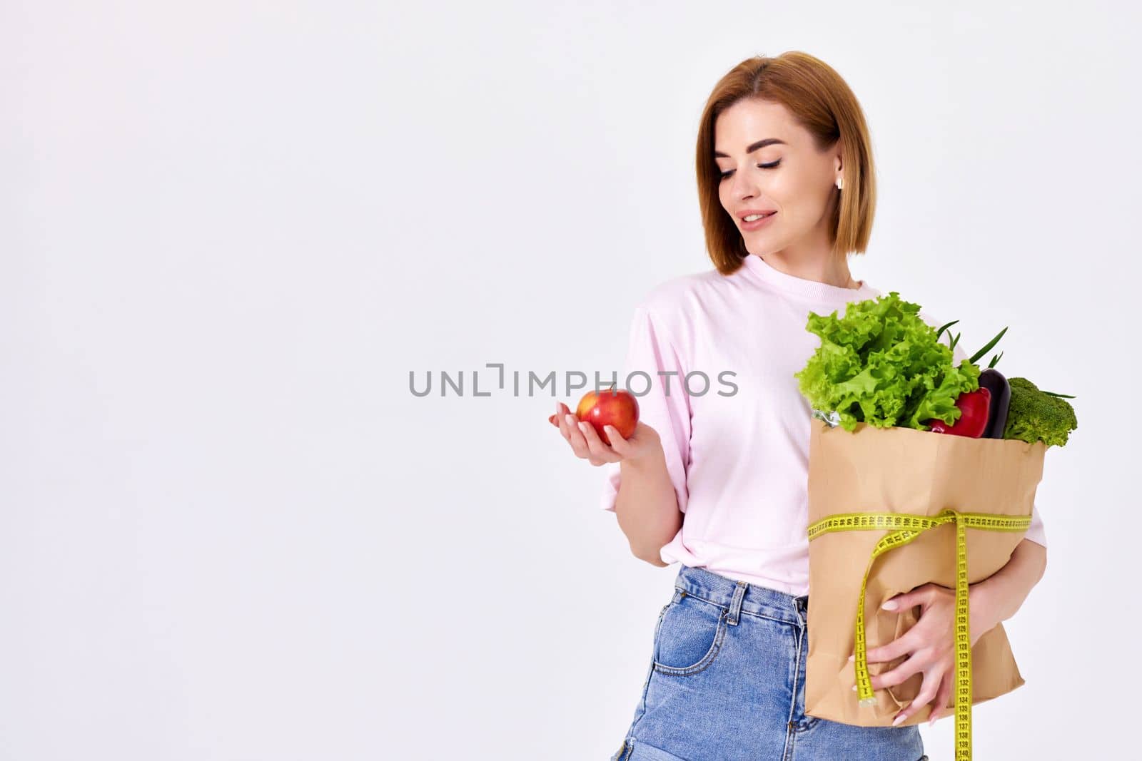 young caucasian woman in pink t-shirt hold paper bag with groceries and with measuring tape and red apple on white background. copy space