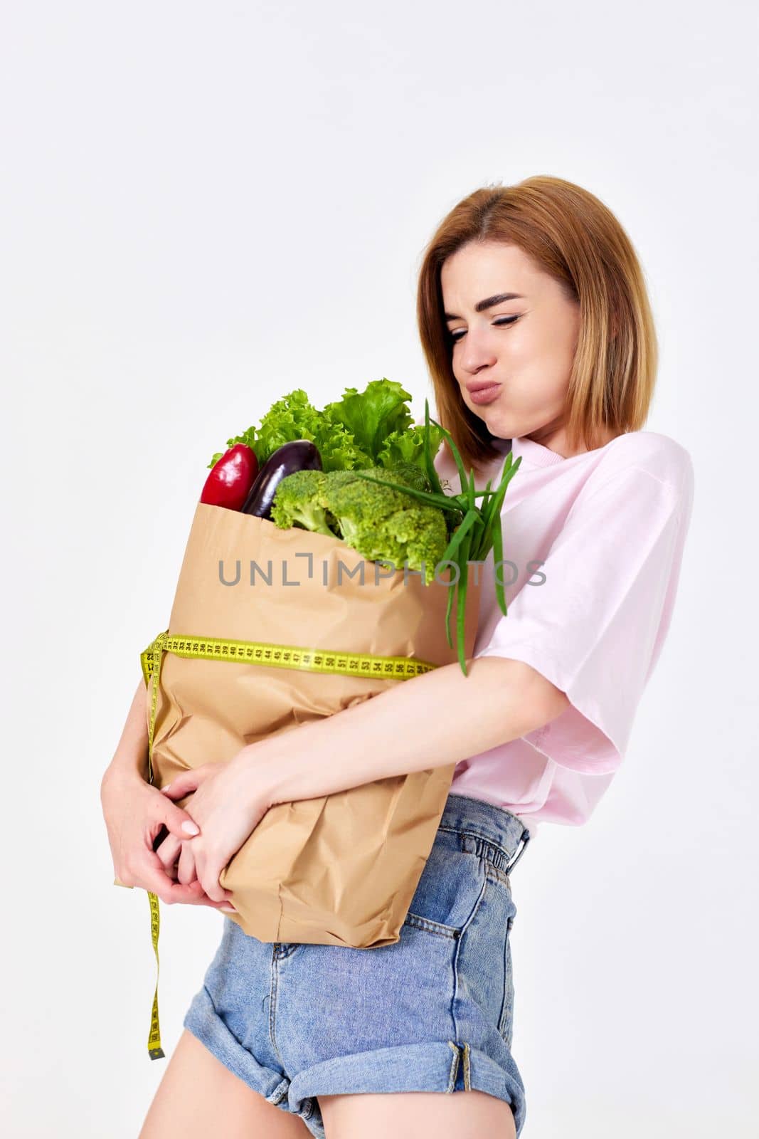 funny young woman holding paper bag with vegetables and measuring tape on white background. healthy food