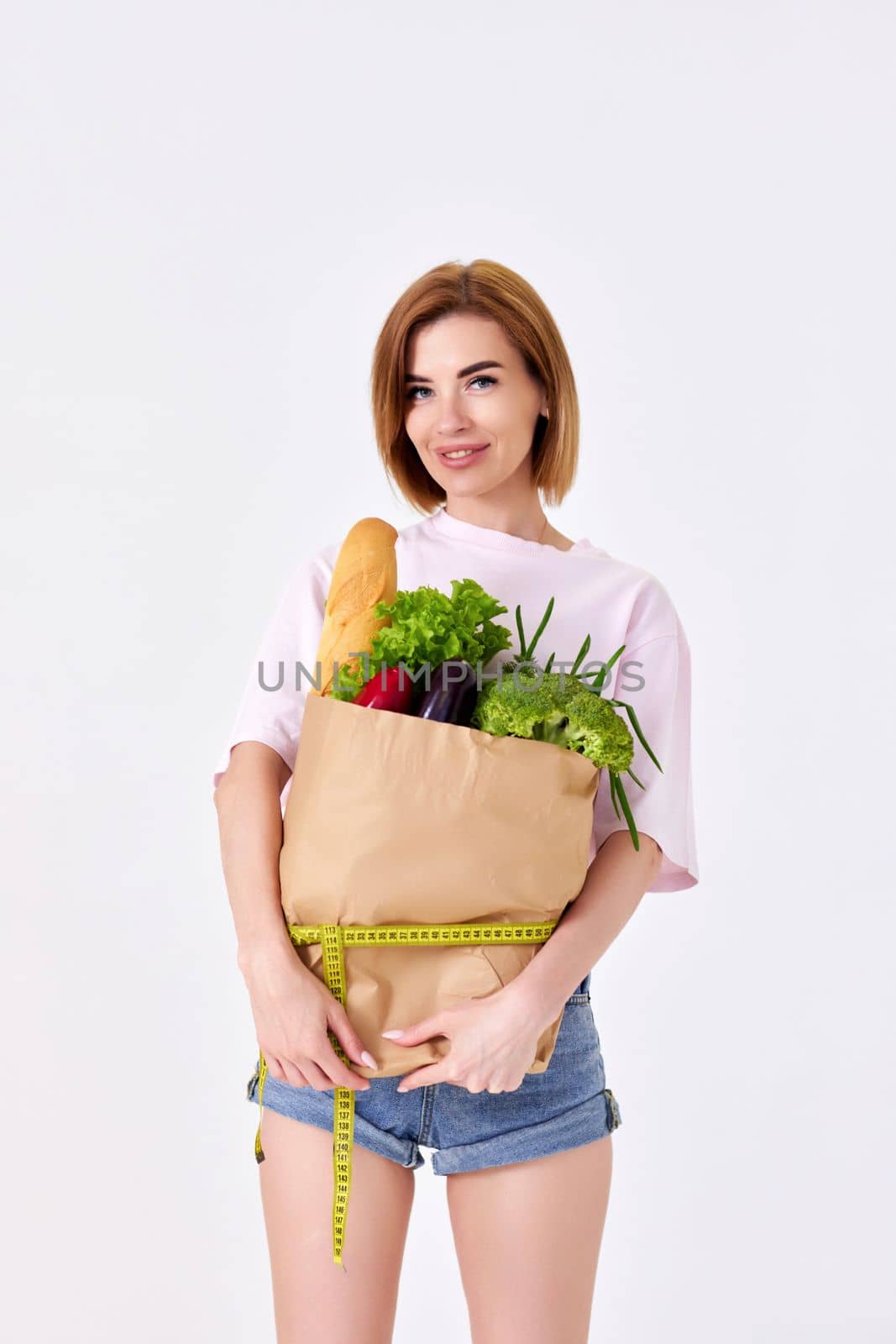 sporty slim woman holding a shopping bag full of groceries and measuring tape on white background.